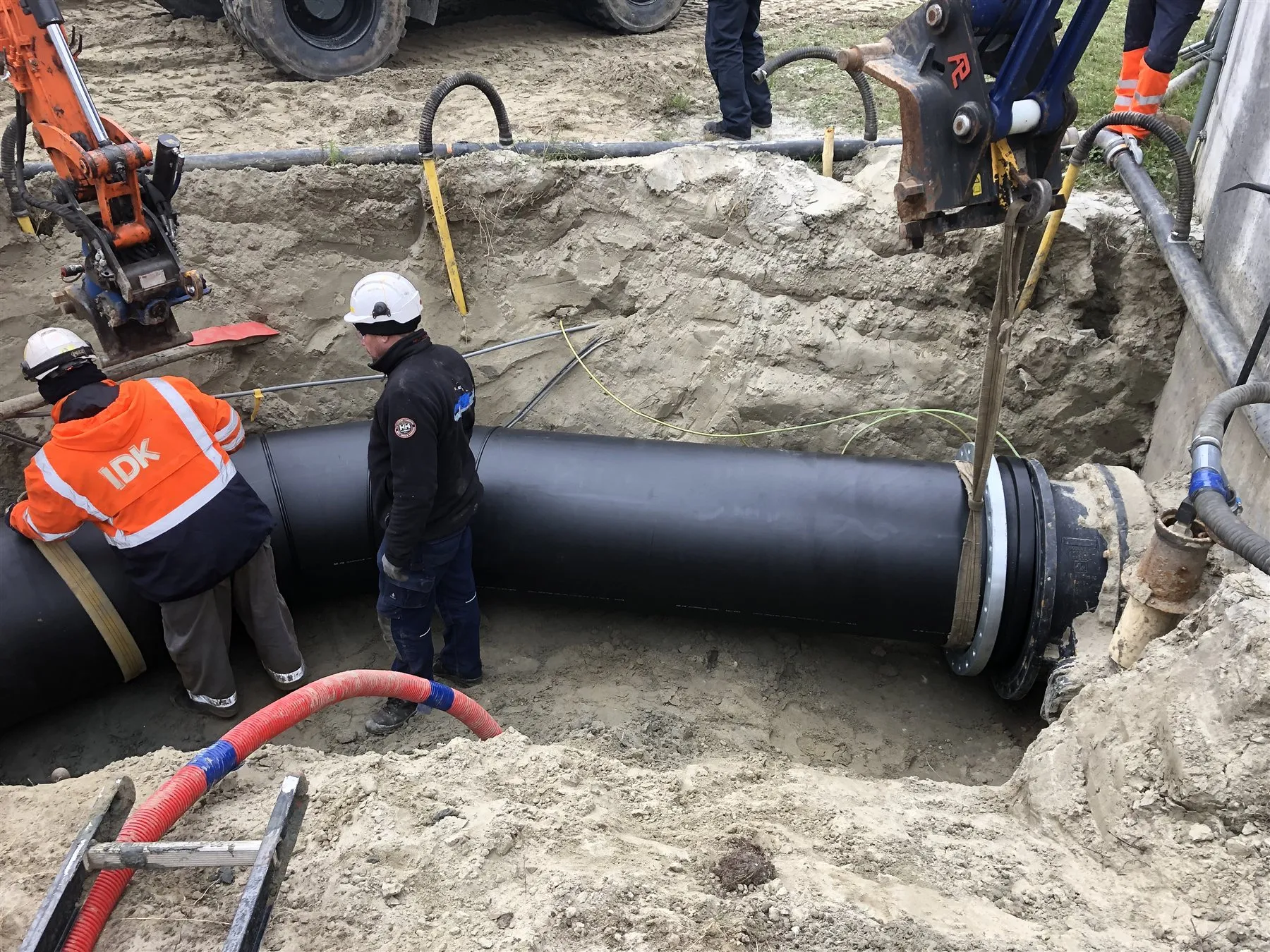 IDK-monteurs laten een grote HDPE-leiding met flens in de sleuf zakken.